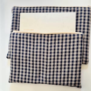 May include: A navy blue and white plaid laptop sleeve with a beige zipper and a white laptop inside. The laptop has the brand name "SAMSUNG" printed on it. The sleeve is made of a soft, quilted fabric.