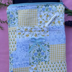 Könnte beinhalten: Eine gesteppte Tablet-Hülle mit einem Patchwork-Design aus Blumen- und Vichy-Mustern in Blau, Gelb und Weiß. Eine hellblaue Schleife verziert die Vordertasche. Ein weißer Stift ist in der Tasche. Der obere Rand hat einen hellblauen Reißverschluss.