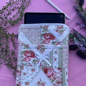 以下が含まれることがあります： 赤バラと緑の葉が白い背景に描かれた花柄のキルトタブレットスリーブ。スリーブには白いジッパーと白いリボンが付いています。白いスタイラスとタブレットが見えます。