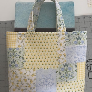 Puede incluir: Un bolso tote acolchado con un diseño de patchwork. El bolso presenta una variedad de telas estampadas florales en amarillo, azul y blanco. Las asas también están acolchadas con un estampado floral. El bolso es rectangular y parece hecho a mano.