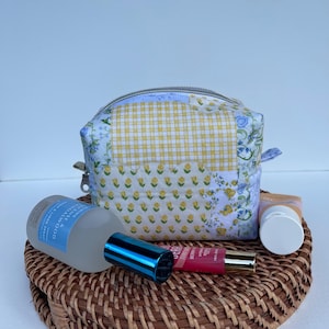 May include: A small, quilted cosmetic bag with a patchwork design featuring yellow gingham, floral, and blue patterns. The bag is open, revealing a silver zip. A bottle of sandalwood spray, a tube of lip gloss, and a small tube of lotion rest on a woven tray.