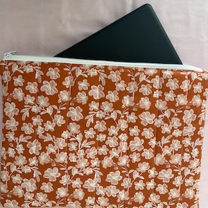 May include: A quilted fabric laptop sleeve with a floral pattern in white on a rust-colored background. The sleeve has a white zipper and holds a black tablet. The sleeve is designed to protect a laptop or tablet.