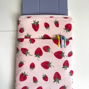 May include: A pink tablet sleeve with a strawberry pattern. The sleeve has a front pocket with three pens and a zipper closure. A blue tablet is partially visible inside the sleeve. The strawberries are red with green leaves and are scattered across the pink fabric.