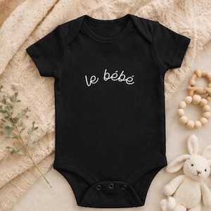 May include: A black baby bodysuit with the French words "le bébé" in white script. The short-sleeved bodysuit has poppers at the bottom. A wooden teething ring and a stuffed bunny are next to the bodysuit.