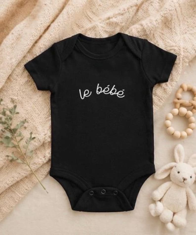 May include: A black baby onesie with the French phrase "le b&eacute;b&eacute;" in white script. The onesie is short-sleeved and has snaps at the bottom. A wooden teething ring and a stuffed bunny are next to the onesie.