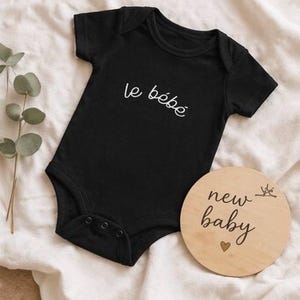 May include: Black baby onesie with the text "le b&eacute;b&eacute;" in white script. A round wooden sign with the words "new baby" and a heart is next to the onesie. The onesie has three snaps at the bottom.