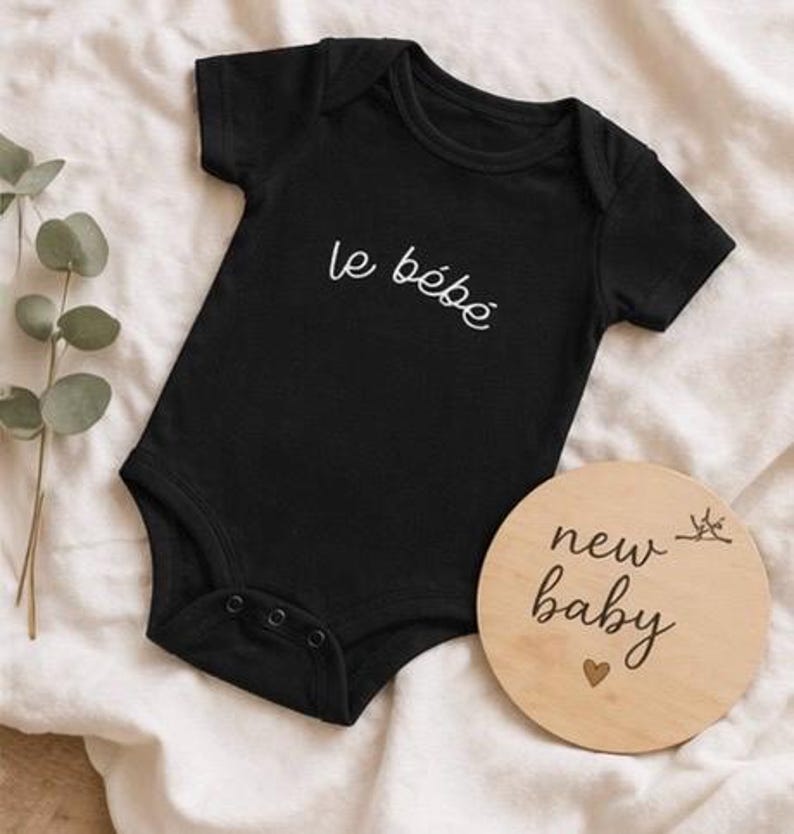 May include: Black baby onesie with the text "le b&eacute;b&eacute;" in white script. A round wooden sign with the words "new baby" and a heart is next to the onesie. The onesie has three snaps at the bottom.