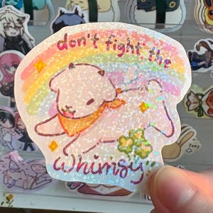 Könnte beinhalten: Holografischer Aufkleber mit einem Cartoon-Schaf, Regenbogen und dem Text "don't fight the whimsy". Das Schaf trägt ein orangefarbenes Halstuch, mit Sternen und Kleeblättern. Der Aufkleber hat einen Glitzereffekt.