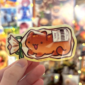 May include: A sticker depicting a cartoon cat-shaped loaf of bread in a clear bag tied with a green bow. The bread is brown with a smiling face and the text "kitty bread!" on a label.