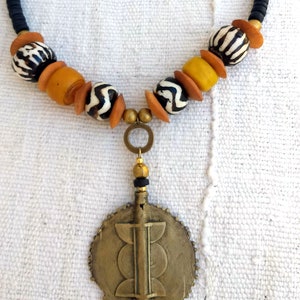 May include: A black and gold beaded necklace with a large, round, gold pendant. The pendant has a geometric design and is attached to the necklace with a gold chain.