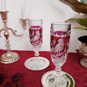 Pode incluir: Duas taças de champanhe de cristal com um desenho de pássaro, sobre bases de prata. As taças têm um interior magenta e estão sobre uma toalha de mesa vermelha. Um castiçal de prata e uma planta estão no fundo.