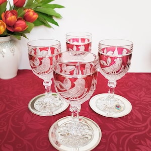 May include: Four crystal wine glasses with a red interior and etched bird design. The glasses sit on silver coasters, arranged on a red patterned tablecloth. A white vase with orange tulips is in the background.