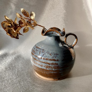 Może przedstawiać: Mały, ręcznie robiony ceramiczny wazon z jasnoniebieskim i brązowym szkliwem, z falbanką i zakrzywionym uchwytem. Złota metalowa róża jest włożona do wazonu, tworząc dekoracyjną kompozycję kwiatową.