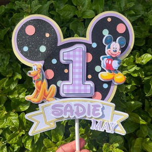 May include: A vibrant cake topper with a Mickey Mouse theme. It features a black Mickey Mouse head silhouette, the number 1, Pluto, and Mickey Mouse. The banner displays the name "Sadie May".