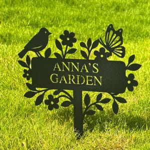 Puede incluir: Letrero de jardín de metal negro con el texto "ANNA'S GARDEN". El letrero presenta un pájaro, una mariposa, flores y hojas. El letrero está colocado en hierba verde.