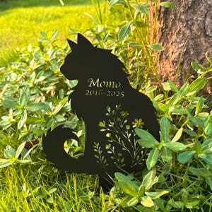 May include: Black metal cat silhouette garden stake with the name "Momo" and the years "2016-2025" inscribed. The cat's body features a floral design. The stake is placed in a bed of green grass and foliage.