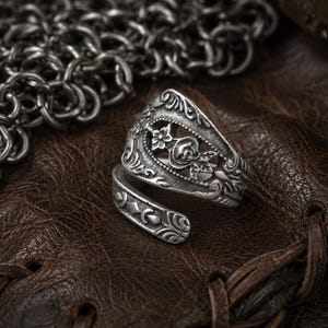 May include: A silver-toned metal ring featuring an open-ended wrap design. The surface of the ring displays an intricate embossed pattern with floral motifs, including a central rose, and decorative scrollwork along the band's edges.