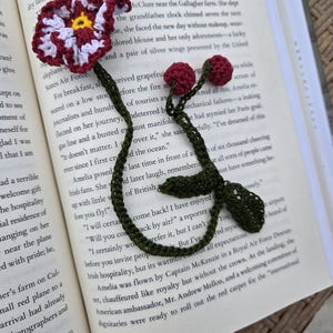 May include: Crocheted flower bookmark with a burgundy and white flower, yellow center, and green stem and leaves. The bookmark is placed in an open book.