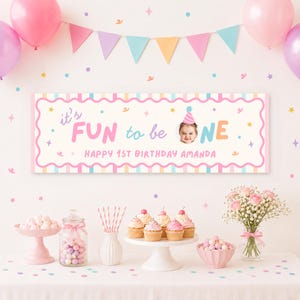 May include: A rectangular birthday banner with a scalloped border featuring the text it's FUN to be ONE and HAPPY 1ST BIRTHDAY AMANDA, including a circular photo of a child wearing a party hat, set against a backdrop of balloons and cupcakes.