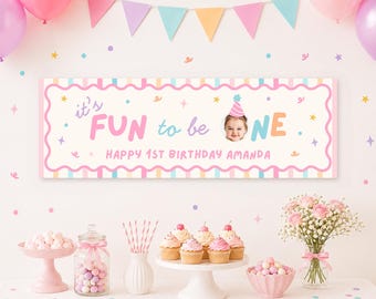 May include: A rectangular birthday banner with a scalloped border featuring the text it's FUN to be ONE and HAPPY 1ST BIRTHDAY AMANDA, including a circular photo of a child wearing a party hat, set against a backdrop of balloons and cupcakes.