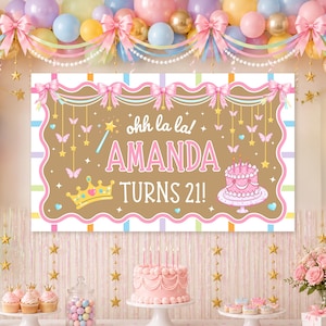 May include: A rectangular birthday banner with a tan background, pink scalloped border, and pink bows. It features the text ohh la la! AMANDA TURNS 21!, a gold crown, a pink cake, and hanging butterflies and stars against a striped backdrop.
