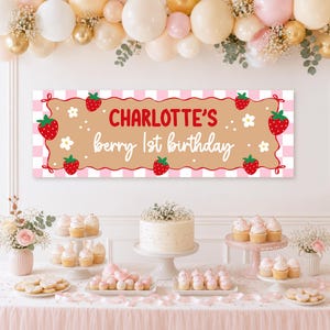 May include: A rectangular party banner with a light pink and white checkered border, featuring a tan center panel decorated with red strawberries and white flowers. The text reads Charlotte's berry 1st birthday in red and white script.