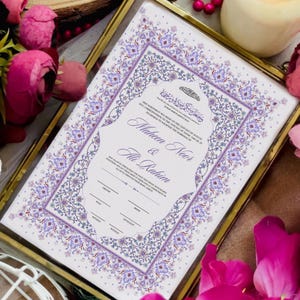 Puede incluir: Un certificado de boda enmarcado con un borde floral decorativo en tonos morados y blancos. El certificado incluye los nombres "Maheen Noor & Ali Rahim" y texto en ingl&eacute;s y &aacute;rabe. El marco es dorado.