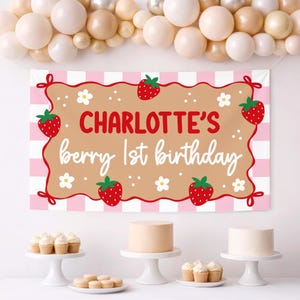 May include: A rectangular party banner with a pink and white checkered border and a tan center featuring red strawberries, white flowers, and the text Charlotte's berry 1st birthday in red and white script and block lettering fonts.
