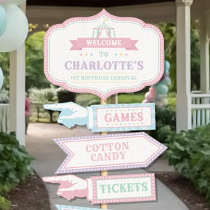 Könnte beinhalten: Ein Satz Schilder im Karneval-Stil für eine Geburtstagsfeier. Das Hauptschild lautet "WELCOME TO CHARLOTTE'S 1ST BIRTHAY CARNIVAL". Weitere Schilder weisen auf "SPIELE", "ZUCKERWATTE" und "TICKETS" hin. Die Schilder sind pastellrosa, blau und weiß.
