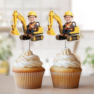 May include: Two cupcakes topped with white frosting and gold sprinkles. Each cupcake has a decorative topper featuring a cartoon image of a child in a yellow excavator. The excavator is yellow and black.