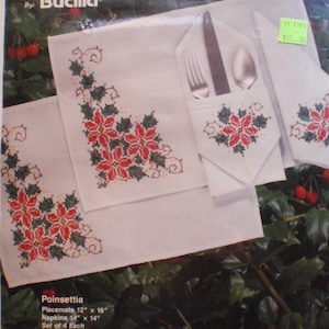 May include: A Bucilla Gallery of Stitches kit featuring a poinsettia design. The kit includes four pieces: two 12" x 16" placemats and two 14" x 14" napkins. The white fabric is adorned with red and green cross-stitch poinsettias.
