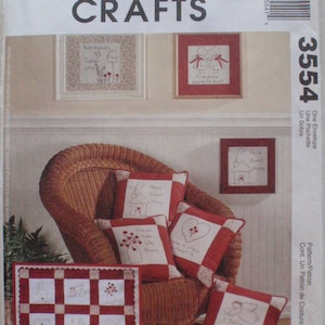 Op de afbeelding: McCall's Crafts patroonpakket met kruissteekontwerpen. De afbeelding toont ingelijste kruissteekkunst en rood-witte kussens met geborduurde ontwerpen. De ontwerpen omvatten huizen, harten, engelen en bloemmotieven. Het pakketnummer is 3554.