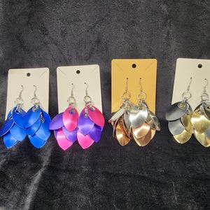 May include: Four pairs of dangle earrings with five layered metal leaves. The earrings are displayed on white and brown card stock. The leaves are in blue, pink, silver, and gold.