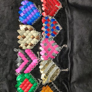 May include: A collection of handmade chainmaille butterfly pendants in various colors, including blue, red, silver, pink, and green. Each pendant is crafted with small metal scales and connected with a chain. The pendants are displayed on a black surface.