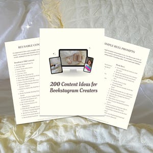 May include: Three cream-colored sheets of paper with text, including "200 Content Ideas for Bookstagram Creators." The image shows a computer monitor and two mobile devices with images. The papers are on a white and yellow bedspread.