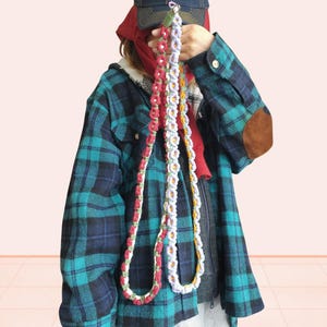 Puede incluir: Tres cordones de ganchillo coloridos hechos a mano con diseños de flores. Un cordón es rojo y blanco, otro es blanco y morado, y el tercero es blanco y amarillo. Los cordones se sostienen contra una persona que lleva una camisa a cuadros.