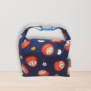 May include: A navy blue bag featuring a Daruma doll pattern in red and white. The bag has a blue handle with a blue clasp. The Daruma dolls are a symbol of good luck in Japan. The bag is made of fabric.