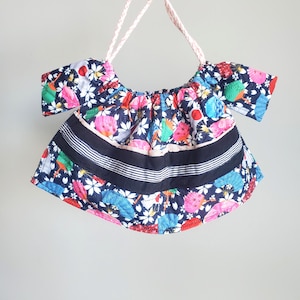 May include: A colorful, off-the-shoulder top with a floral and bird pattern. The top features a navy blue background with white flowers, pink birds, and other colorful accents. It has a black band with white stripes and a pink rope for hanging.