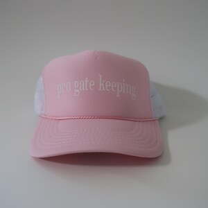 May include: A pink trucker hat with white mesh backing. The hat features the phrase "pro gate keeping" in white lettering across the front. The hat has a curved brim and a pink rope detail above the brim.