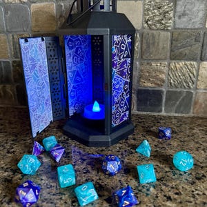May include: A black metal lantern with a blue and white patterned glass insert. The lantern is open and a small blue light is visible inside. There are 10 blue and teal dice scattered on a brown surface.
