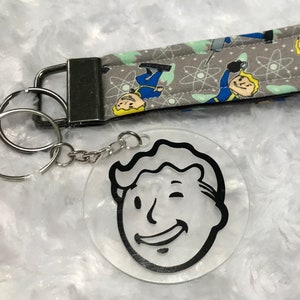 May include: A keychain with a clear, circular charm displaying a black and white cartoon face. The keychain is connected to a grey lanyard patterned with cartoon figures and atoms. The lanyard has a metal clasp and key rings. The design is reminiscent of a video game.