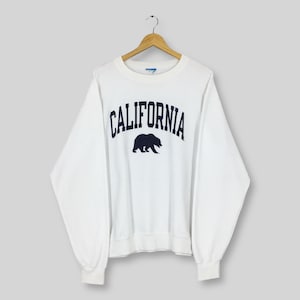 Könnte beinhalten: Weißes Sweatshirt mit dem Wort "CALIFORNIA" in Marineblau, das sich bogenförmig über der Brust wölbt, über einer marineblauen Bärensilhouette. Das Sweatshirt hängt an einem Holzkleiderbügel.