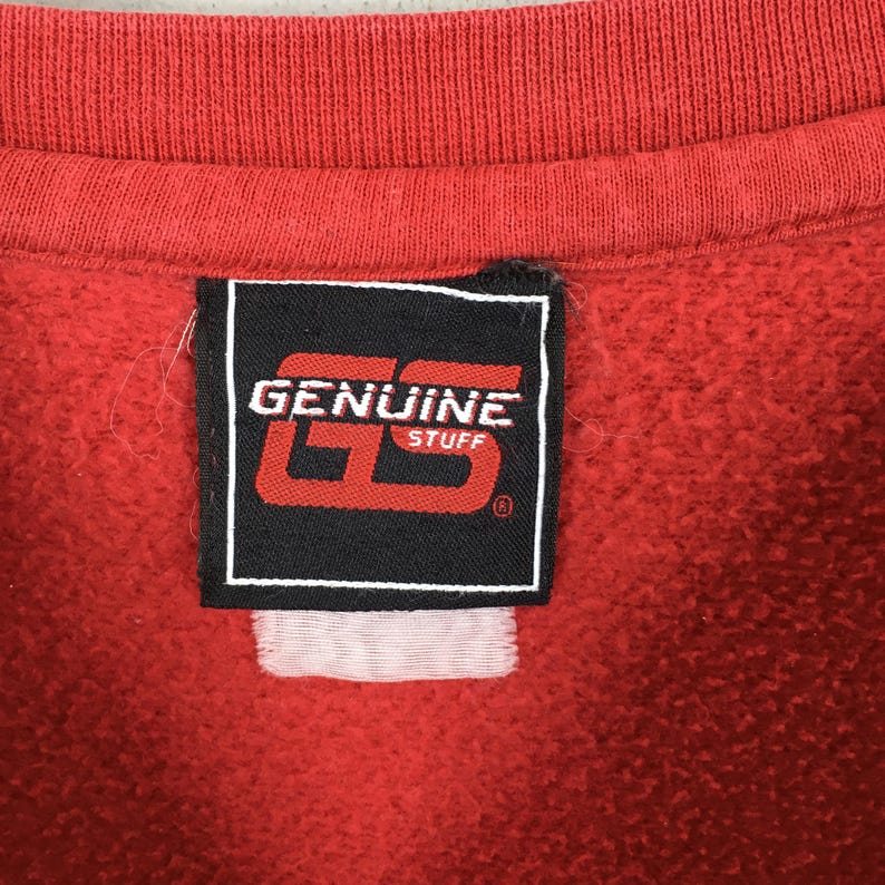 May include: A red sweatshirt featuring a black rectangular fabric label sewn onto the interior neckline. The label displays the white text GENUINE STUFF and a large red GS logo. A small, blank white fabric tag is positioned directly below the label.