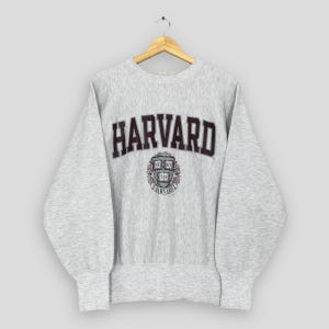 以下が含まれることがあります： 長袖のライトグレーのハーバードスウェットシャツ。「HARVARD」の文字が胸にダークマルーン色でプリントされています。その下には、「VERITAS」と「HARVARD」の文字がダークマルーン色で書かれた円形エンブレムがあります。