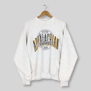 Könnte beinhalten: Ein hellgrau meliertes Sweatshirt mit Rundhalsausschnitt und dem Schriftzug APPALACHIAN in großen schwarzen Buchstaben mit gelber Umrandung. Hinter dem Text befindet sich ein kreisförmiges Siegel mit APPALACHIAN STATE UNIVERSITY und 1899.