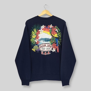May include: Navy blue sweatshirt with a vibrant graphic of a parrot, waves, and tropical flowers. The design includes the text "RON JON SURF SHOP U.S.A." The sweatshirt is displayed on a wooden hanger against a light background.