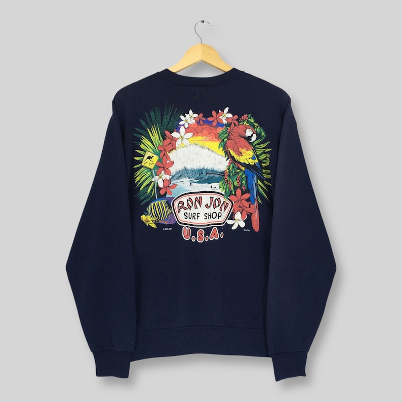 May include: Navy blue sweatshirt with a vibrant graphic of a parrot, waves, and tropical flowers. The design includes the text "RON JON SURF SHOP U.S.A." The sweatshirt is displayed on a wooden hanger against a light background.