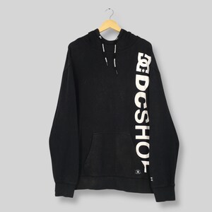 May include: Black hooded sweatshirt with a kangaroo pocket and white drawstring. The right sleeve features the white text "DC SHOE" vertically. The sweatshirt is hanging on a wooden hanger.