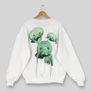 May include: White sweatshirt featuring a detailed illustration of several manatees in shades of green. The manatees are clustered together, creating a unique design on the back of the garment. The sweatshirt has long sleeves and a crew neck.