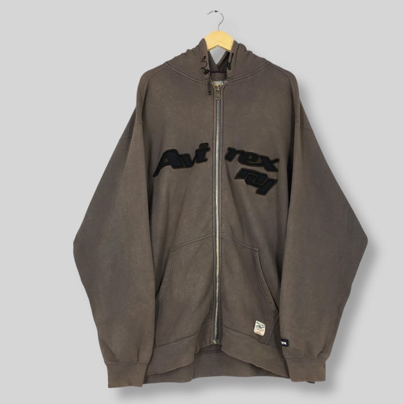 Puede incluir: Una sudadera con capucha marr&oacute;n con cremallera y el texto "Avt Rex Foy" en negro, bordado en la parte delantera. La sudadera tiene una capucha con cord&oacute;n, un bolsillo delantero y una peque&ntilde;a etiqueta en la parte inferior. La prenda est&aacute; colgada en una percha de madera.